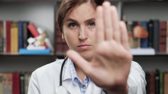 医生显示停止。穿着白大褂和听诊器的兴奋女医生在办公室看着相机举起她的手，用手掌显示停车标志。爆头