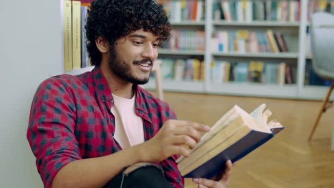 读书。英俊聪明的印度或阿拉伯大学或大学男生，坐在靠近书架的图书馆的地板上，阅读有趣的书，准备考试，获