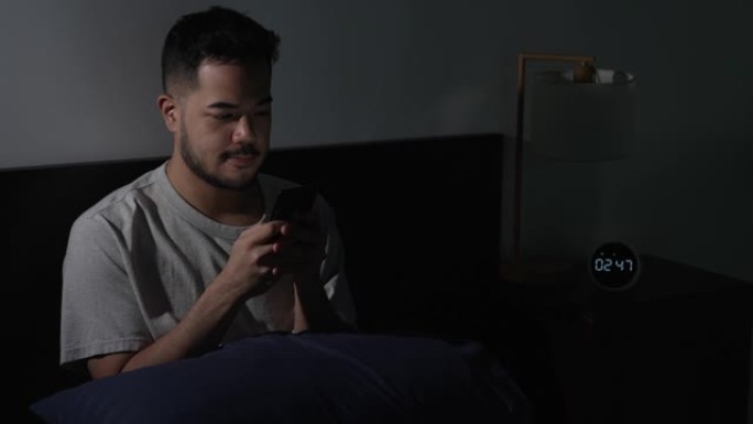 年轻的亚洲男子晚上躺在床上使用智能手机时眼睛酸痛疲惫。