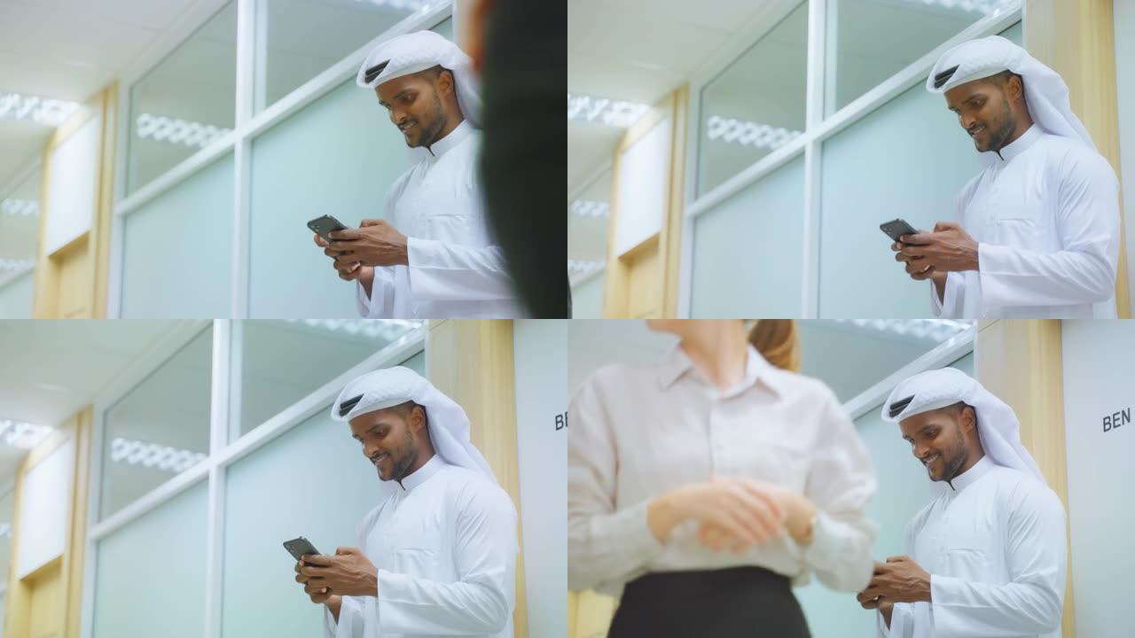 阿拉伯商人站在办公室走廊上使用智能手机。有吸引力的男性员工在室内与同事聊天并在手机上交流对企业工作场