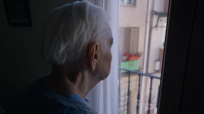 特写-灰色头发皱纹的老妇人站在黑暗中看着窗外。无法辨认的高加索女性高级退休人员欣赏着站在室内的阳光明