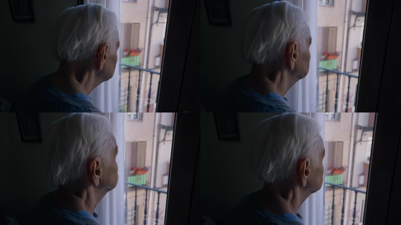特写-灰色头发皱纹的老妇人站在黑暗中看着窗外。无法辨认的高加索女性高级退休人员欣赏着站在室内的阳光明