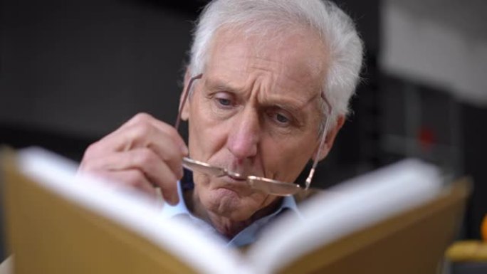 集中老人阅读书籍脱下眼镜翻页的特写正视图。专注的高加索人感兴趣的男性退休人员在家室内享受文学。