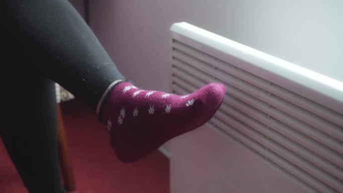 在电暖器的背景上，腿穿着温暖的袜子。寒冷的季节。节约电能。能源危机。可怜的公民。