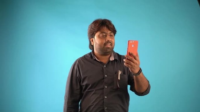 一名穿着黑色衬衫的印度男子站在孤立的蓝色背景下与手机进行视频聊天