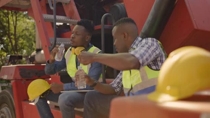 两名非裔美国建筑工人高兴地坐在叉车上喝水，并在工作施工期间与他们交谈。工头与下属友好地提供建议。团队