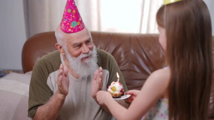 戴着礼帽的微笑老人说话，用燃烧的蜡烛感谢纸杯蛋糕。快乐的高加索祖父的肖像与孙女在家里的客厅庆祝生日。