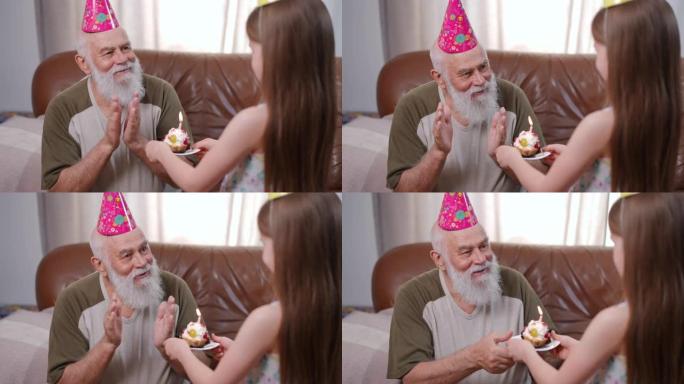 戴着礼帽的微笑老人说话，用燃烧的蜡烛感谢纸杯蛋糕。快乐的高加索祖父的肖像与孙女在家里的客厅庆祝生日。
