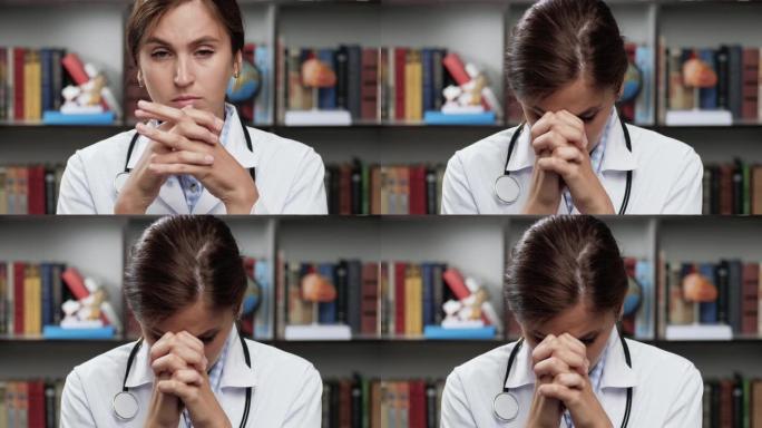医生正在祈祷。穿着白大褂和办公室听诊器的心烦意乱的女医生看着相机，把手举到脸上，开始祈祷。爆头