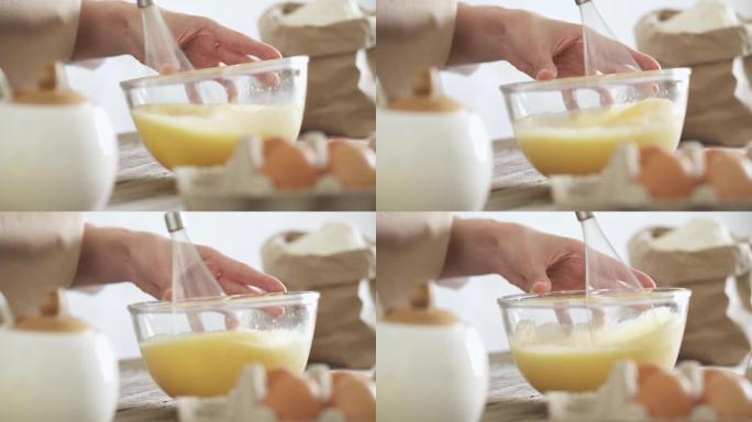 雌性手混合鸡蛋。特写。厨房电动搅拌机在透明碗中敲鸡蛋。鸡蛋托盘