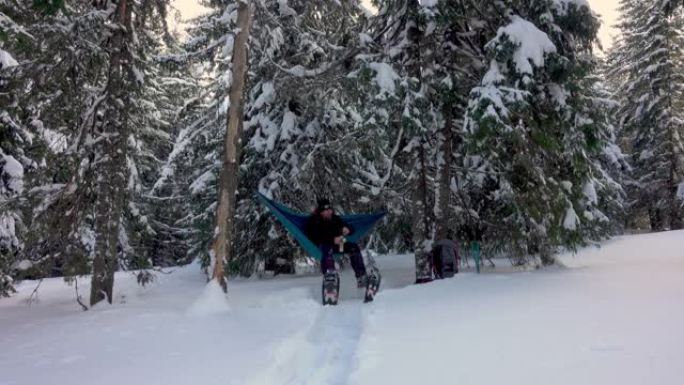 徒步旅行者带着雪鞋喝咖啡，躺在冬季森林的吊床上