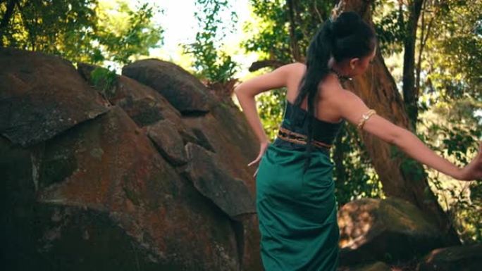 一个穿着传统舞蹈服装的亚洲女人在大石头附近跳舞，背景是棕色的叶子