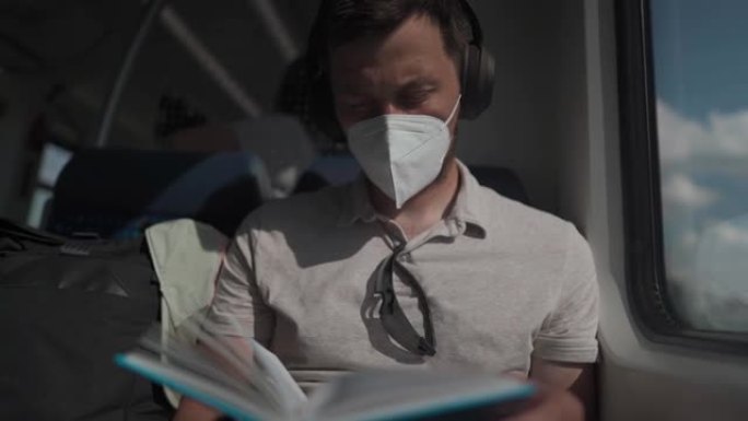 白人男子戴着口罩和耳机坐在火车上，坐在窗户旁看书。男性阅读书籍，并在火车上的耳机上听音乐。主题穿越德