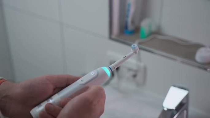 男人的手在浴室背景下打开或关闭带有蓝光的声波白色牙刷。牙齿卫生和洁白的牙齿。电动牙刷。清洁口腔的电动