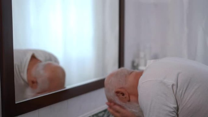 自信的老人洗掉浴室镜子里脸上残留的剃须泡沫。集中的高级高加索退休人员早上在家室内塑造白色长胡须。