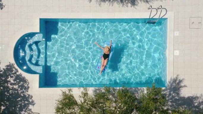 游泳池上的4k镜头，穿着黑色泳衣的放松女人，草帽漂浮在橄榄树下蓝色涟漪水面的充气床垫上。假期，娱乐概