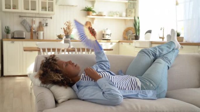 家庭过热，疲惫的非洲裔美国妇女在室内使用手扇躺在沙发上