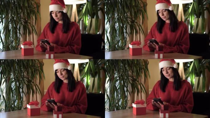 穿着圣诞老人服装的女人坐在家里的桌子旁，手里拿着智能手机，翻阅新闻。使用互联网与亲人交流的概念