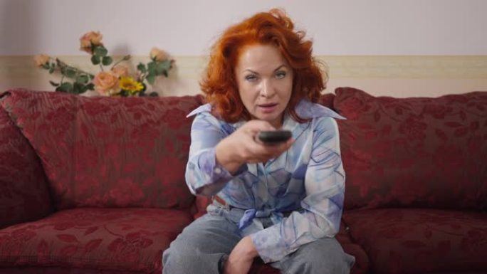 前视图复古女人看着相机选择带遥控器的频道切换。20世纪80年代周末，白人红发女士坐在家里舒适的沙发上