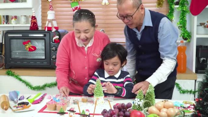 亚洲男孩和祖父母一起制作和装饰圣诞树蛋糕，为圣诞晚会做准备。一家人一起在装饰有装饰品的厨房里度过快乐