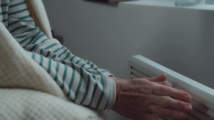 一位肩膀上披着毯子的老妇人用电热器温暖她冰冷的双手。寒冷的季节。家庭预算储蓄。能源危机。可怜的公民。
