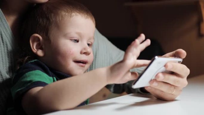 婴儿点击手机屏幕。女人的手握着手机，可爱的2-3岁的蓝眼睛孩子坐在母亲的怀里，在智能手机屏幕上轻拍手