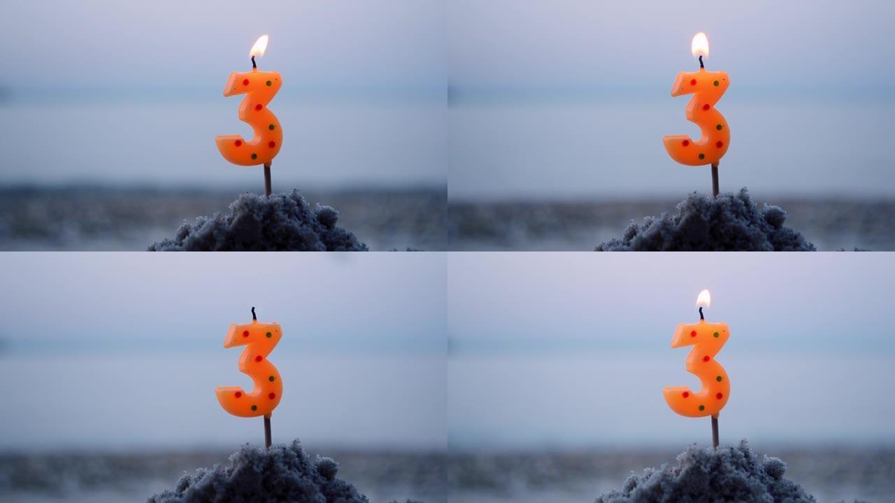 三号蜡烛在棍子上插入沙子和燃烧。模糊海浪背景下的沙子点燃蜡烛。模糊的蓝色棕色背景上的绿色燃烧蜡烛。生