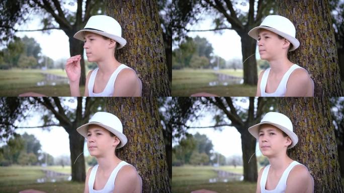 一个戴着帽子的有趣男孩的肖像坐在树下的公园里，在夏日里环顾四周