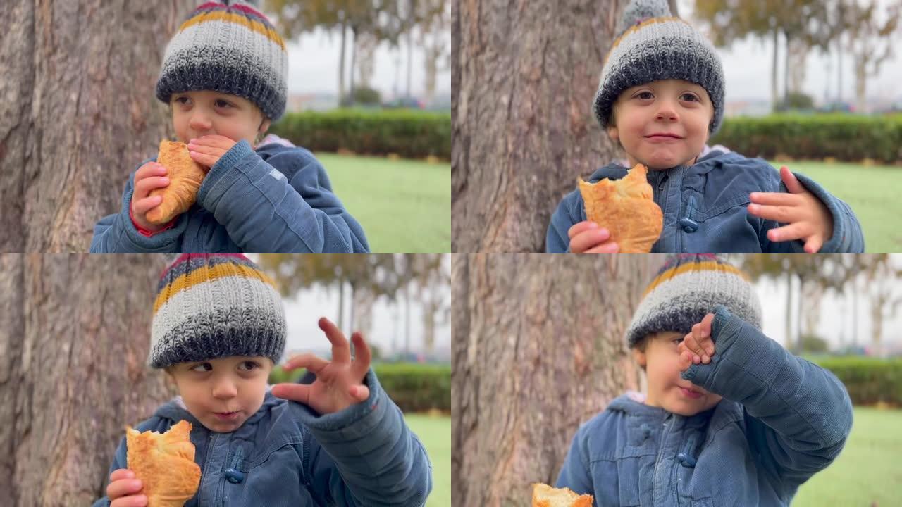 在公园外面吃羊角面包的快乐孩子。小男孩穿着冬装夹克和豆豆。儿童零食碳水化合物食品