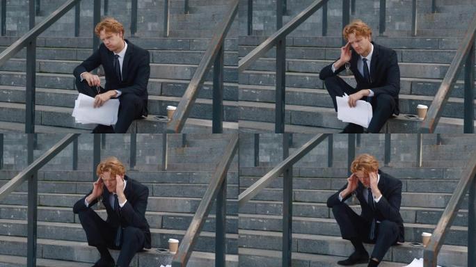 紧张的商人手里拿着商业文件坐在办公楼的台阶上。悲伤过度劳累的红发男人经理，摘下眼镜，把手放在头痛的头
