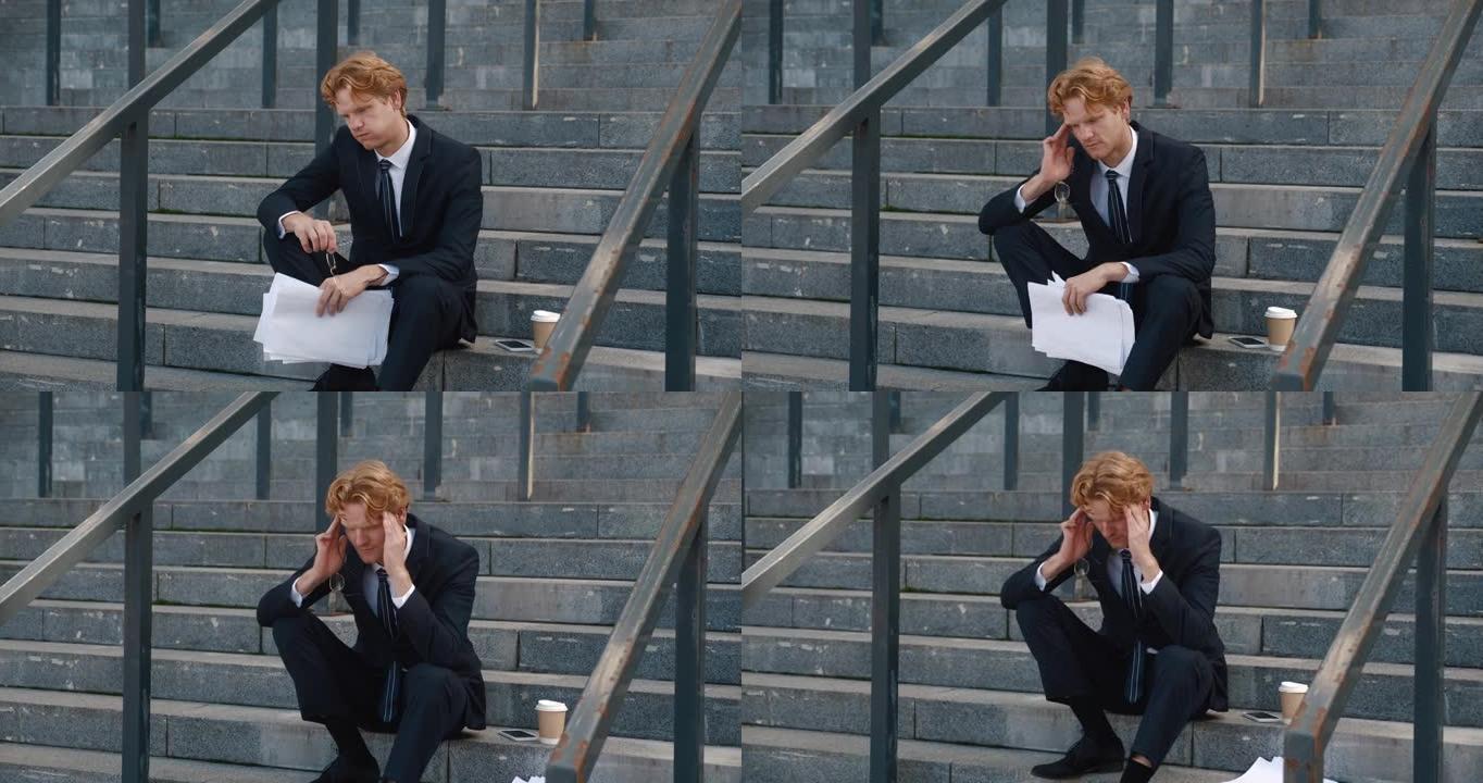 紧张的商人手里拿着商业文件坐在办公楼的台阶上。悲伤过度劳累的红发男人经理，摘下眼镜，把手放在头痛的头