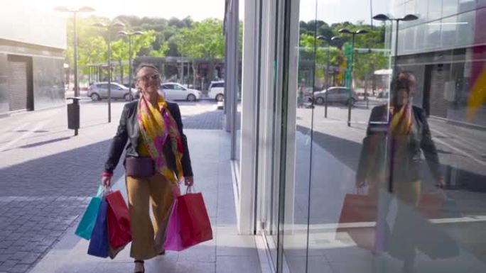 快乐的高级迷人女士带着购物袋走在街上微笑。进入商场。