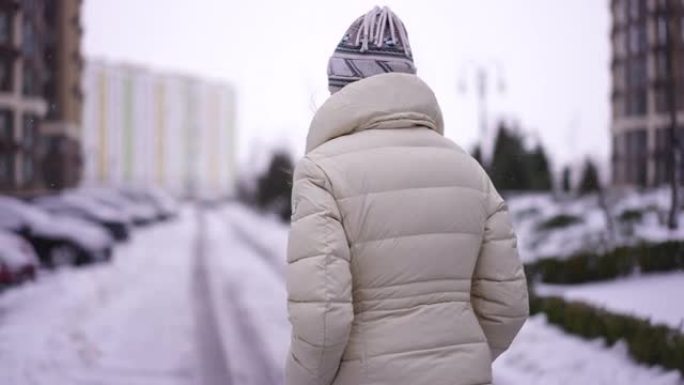 阴天，自信的女人在雪城街上行走的跟踪镜头。现场摄像机跟随穿着白色保暖夹克的苗条白人女士在户外漫步。