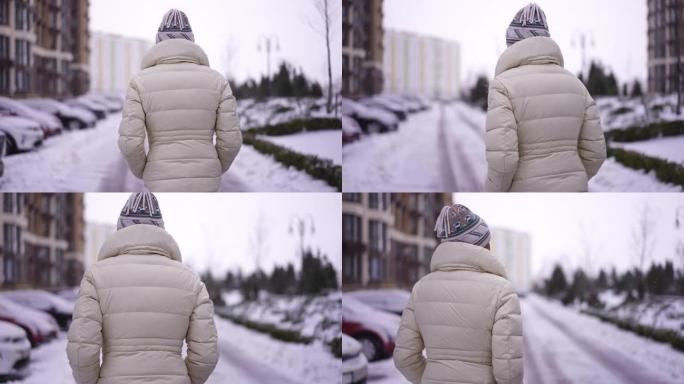阴天，自信的女人在雪城街上行走的跟踪镜头。现场摄像机跟随穿着白色保暖夹克的苗条白人女士在户外漫步。