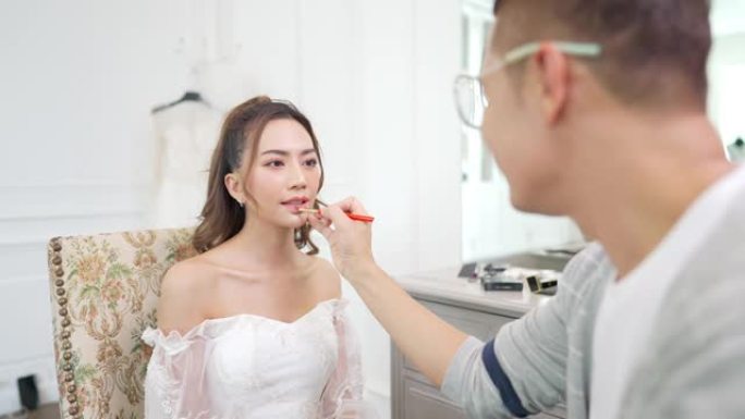 4k亚洲男化妆师为穿着白色婚纱的女性新娘化妆。