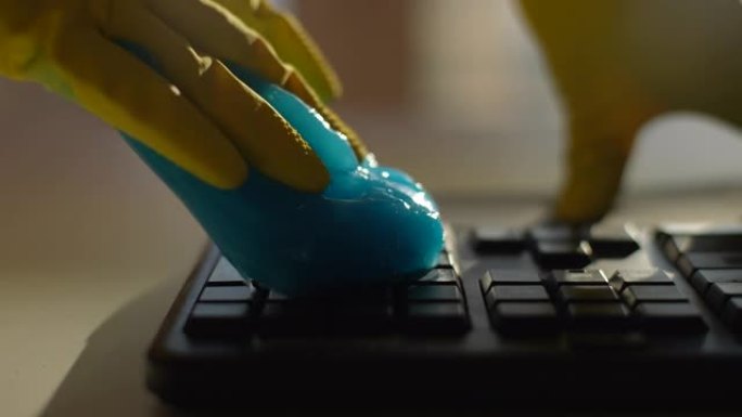 穿着黄色手套的无法识别的年轻人的特写镜头使用蓝色粘液吸收凝胶，慢动作清洁和清除pc键盘上的碎屑。