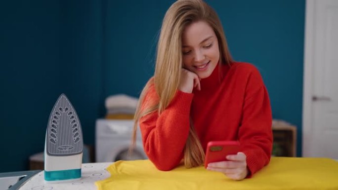 年轻的金发女子在洗衣房用智能手机熨烫衣服