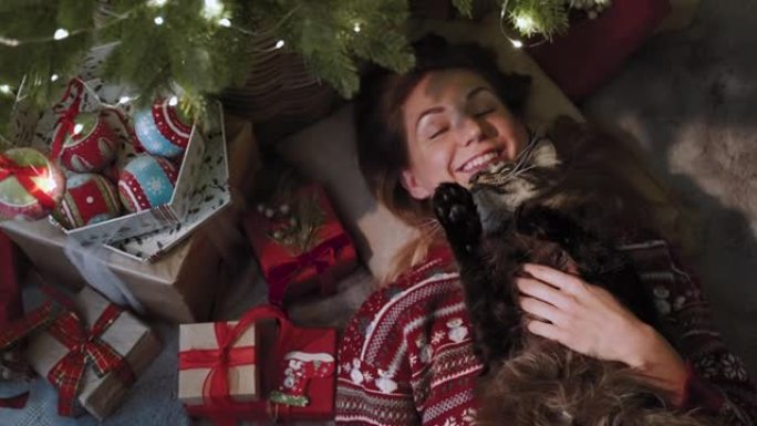一个美丽的女人，在她最喜欢的猫的陪伴下，躺在圣诞树下，有很多礼物。新年庆祝概念。