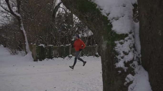 德国冬天，男人在白雪皑皑的小路上慢跑穿过城市的公园，戴着耳机听音乐。运动员在寒冷的天气里戴着耳机锻炼