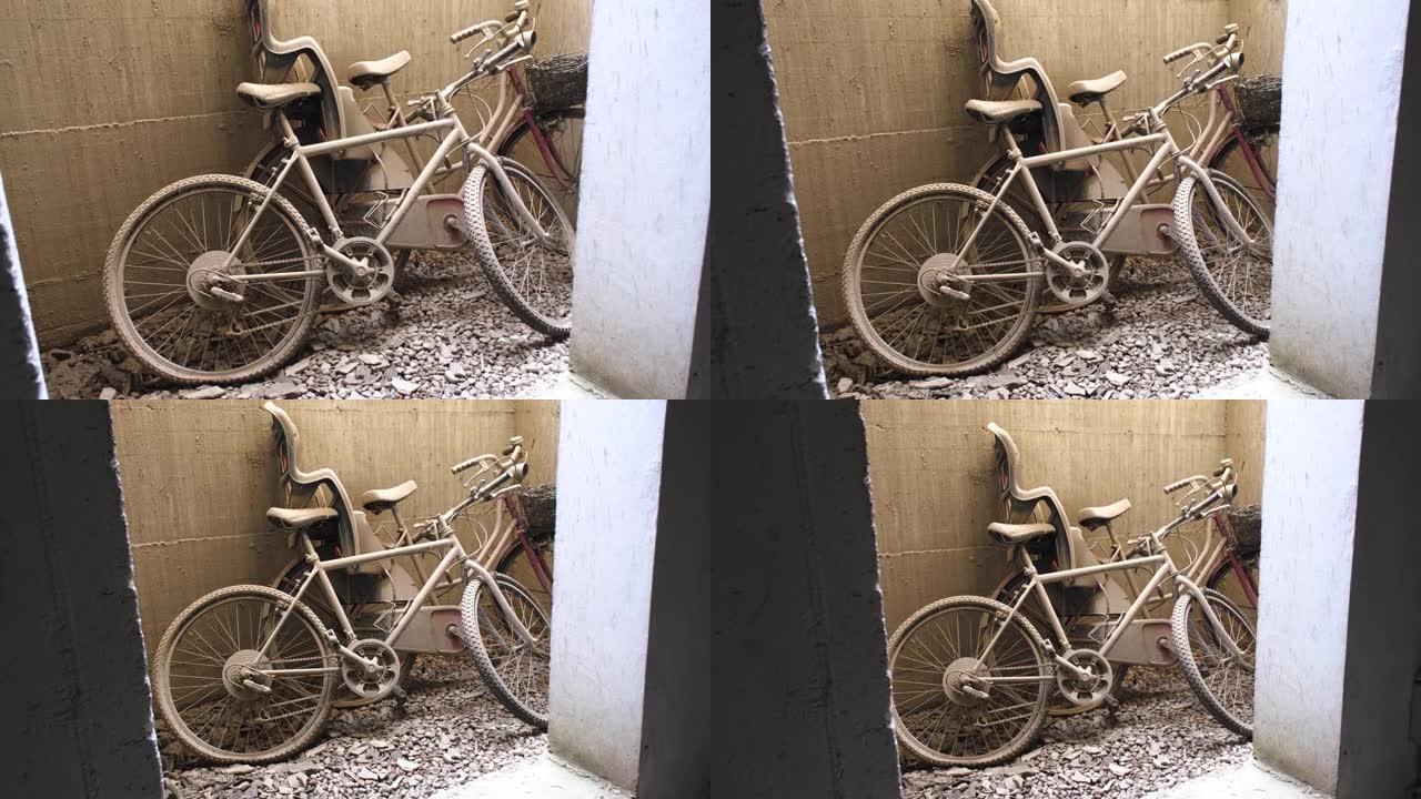 两辆轮胎漏气的废弃自行车靠在沙壁上，在瓦砾覆盖的地面上。儿童座椅，覆盖着泥土