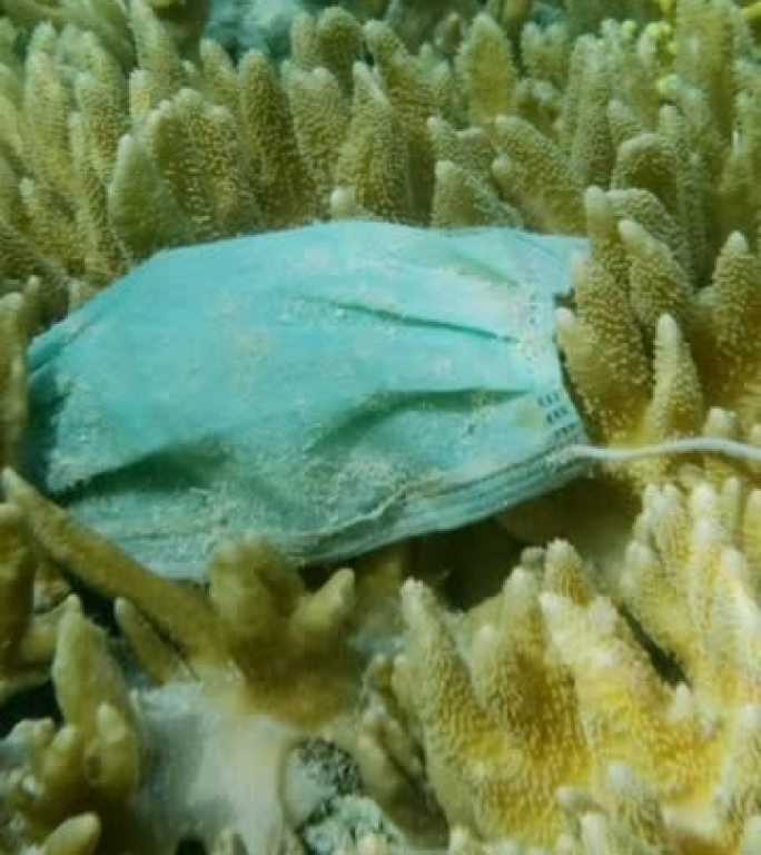 废弃的二手医用口罩的特写镜头位于阳光下美丽的柔软珊瑚的水下。海洋的污染面具。慢动作，摄像机向前移动