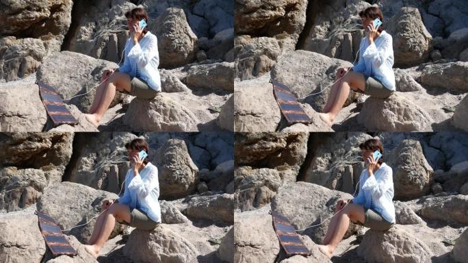 一个旅游女孩坐在山上的石头上，用智能手机说话，智能手机是用太阳能电池板充电的。