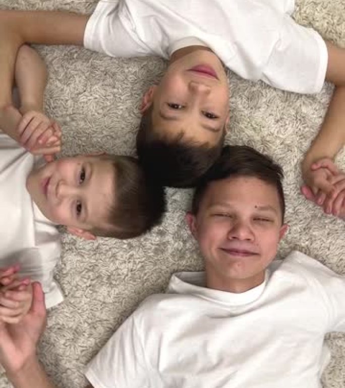 垂直视频兄弟家庭三个朋友躺在上面牵着手微笑快乐快乐一起一群快乐的孩子玩耍的孩子快乐的朋友躺在地毯上看