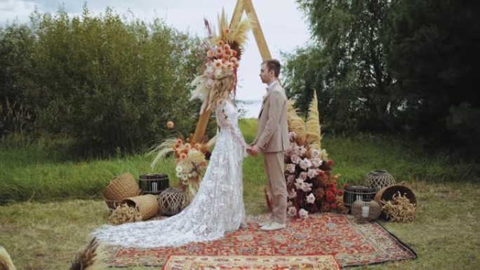 美丽的新婚夫妇站在装饰好的波西米亚婚礼拱门附近牵手，慢动作。