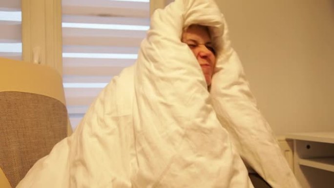 冰冻的女人坐在昏暗的寒冷房间里，用温暖的白色毯子完全包裹身体，摩擦脸颊。室内低温。冬天，寒冷，沮丧，