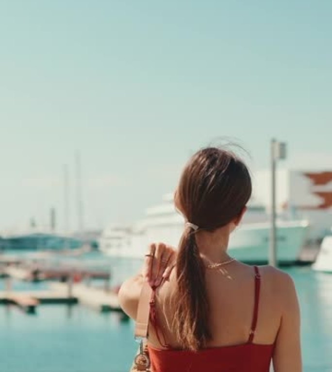 垂直视频: 特写可爱的女人在海港背景上行走。女孩喜欢看游艇和轮船的好天气和美丽的景色。后视图