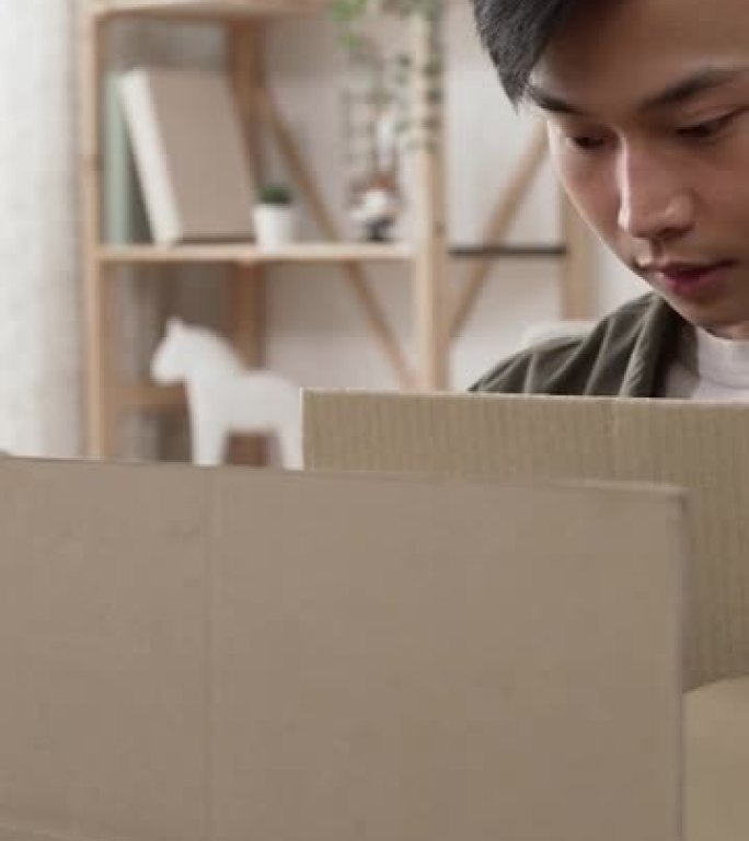垂直视频: 快乐的韩国男顾客坐在沙发上打开包裹纸箱的包装对在家通过邮政运输的赠品包装感到惊讶和满意