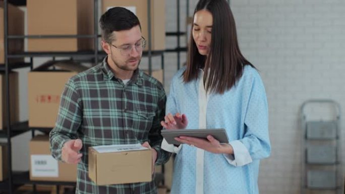 一位使用平板电脑的女性销售经理正在与一位拿着纸板包装的男性工人交谈。后台准备装运包裹的仓库。