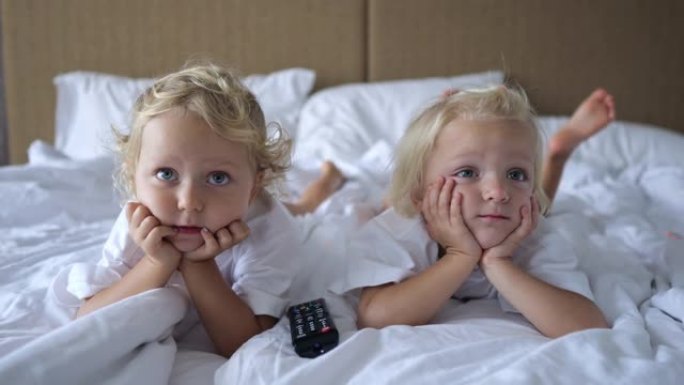 三岁的小姐妹躺在床上，双手放在下巴上，看着电视屏幕。他们仔细观看儿童动画片。