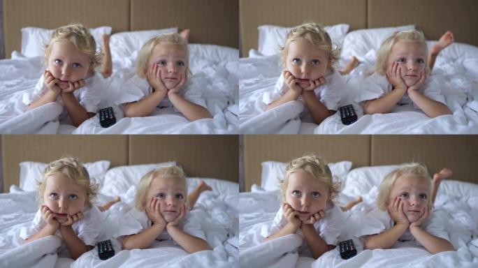 三岁的小姐妹躺在床上，双手放在下巴上，看着电视屏幕。他们仔细观看儿童动画片。